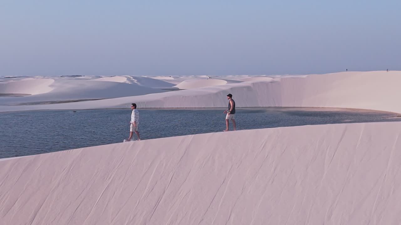 Aerial shot pan left drone view two boys or men over the white sand dunes and crystal clear water lagoons of the Lencois Maranhenses in Maranhao, Brazil. UNESCO World Heritage Site. Establishing Shot.