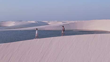 aerial shot pan left drone view two boys or men over the white sand dunes and crystal clear water lagoons of the lencois maranhenses in maranhao, brazil. unesco world heritage site. establishing shot.