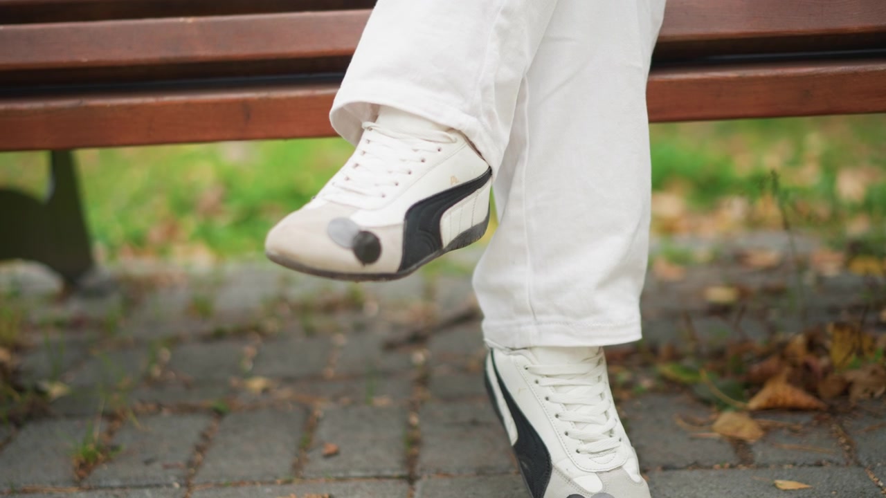 Close-up de menina praticando passos de dança com sapatos e calças brancas de artes marciais no pavimento com folhas caídas. O sujeito realiza movimentos controlados dos pés, demonstrando equilíbrio entre as pernas.