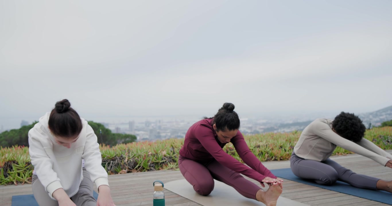 โค้ชหญิงหลากหลายเชื้อชาติกำลังแนะนำการเคลื่อนไหวแบบ lunges ไปยัง seated folds บนเสื่อโยคะที่วางอยู่บนพื้นระเบียง พร้อมขวดสีเขียว