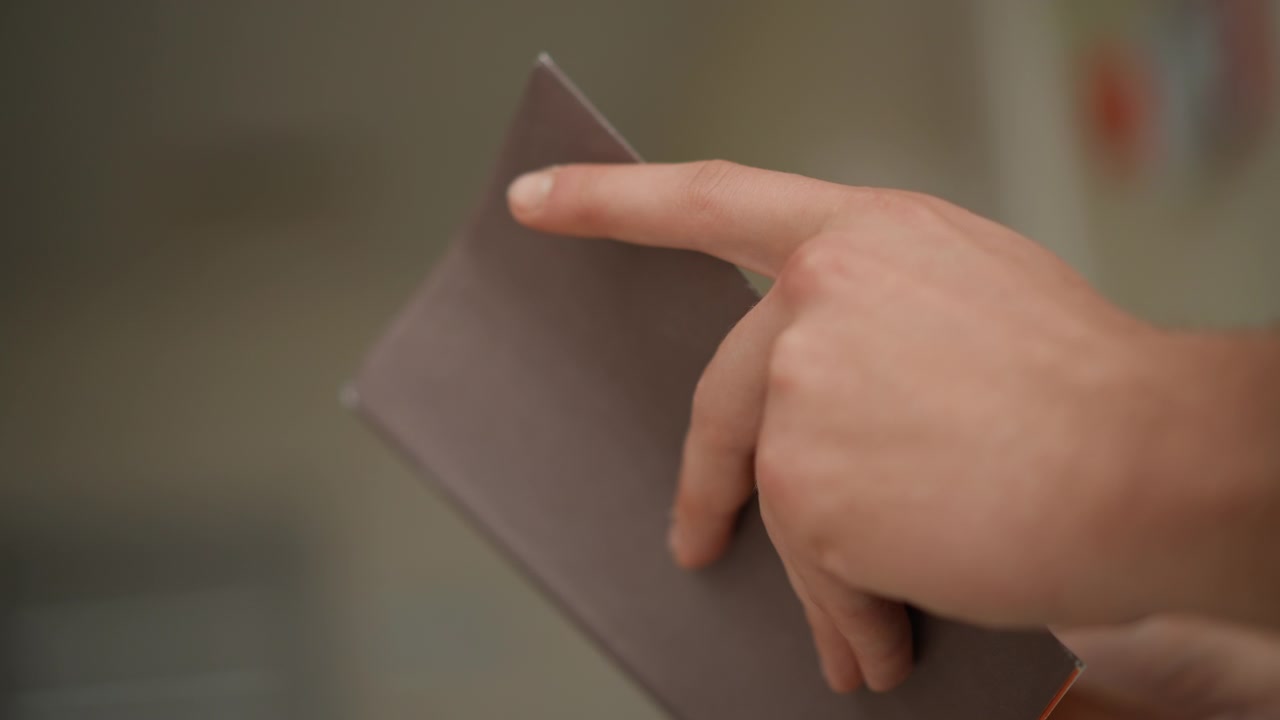 Detailní záběr na ruku držící a ukazující na roh zavřeného zápisníku s měkkým osvětlením a malou hloubkou ostrosti. Scéna se zaměřuje na lidskou interakci s papírnictvím ve vnitřním prostředí.
