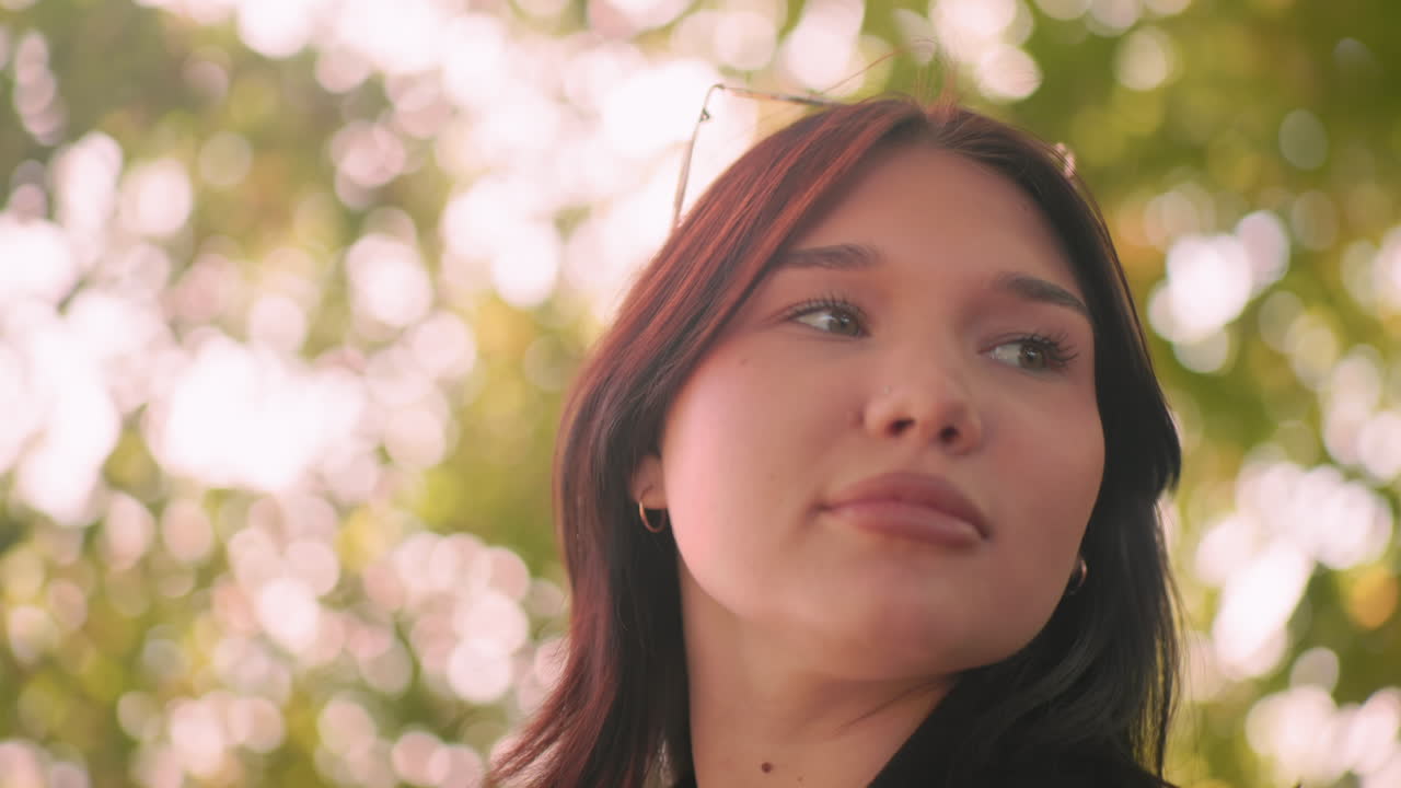 Close up of contemplative girl walking through arboretum, gazing around under autumn canopy, soft bokeh behind, black coat and sweater visible, relaxed pace, quiet mood, gentle sunlight on face