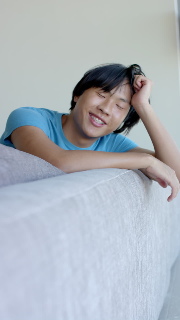 Vertical video: Relaxing on couch, Asian teenage boy smiling and enjoying leisure time at home
