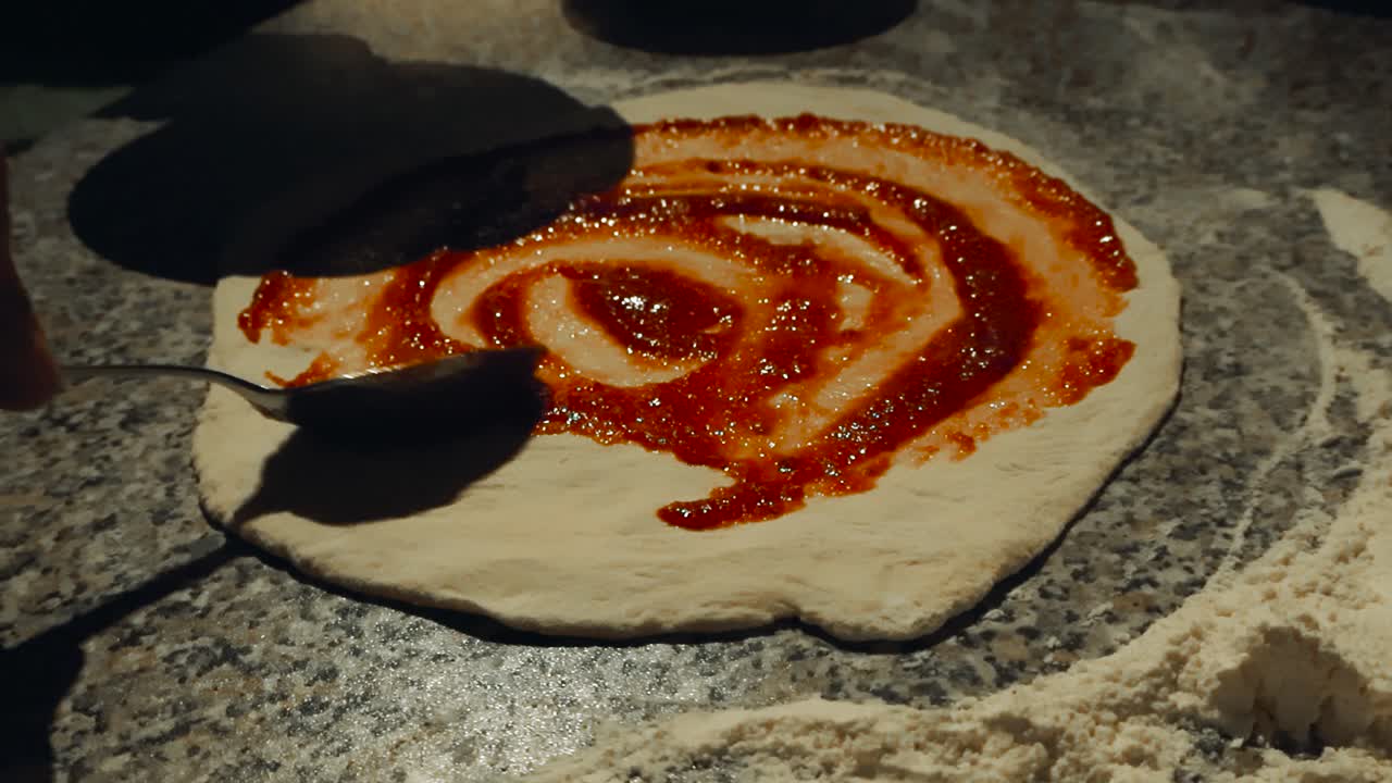 Low angle orbiting of person’s hand gently spreading tomato sauce over raw pizza dough with spoon in close up. Flour dusts around the home kitchen table in shallow depth. Homemade comfort food cooking
