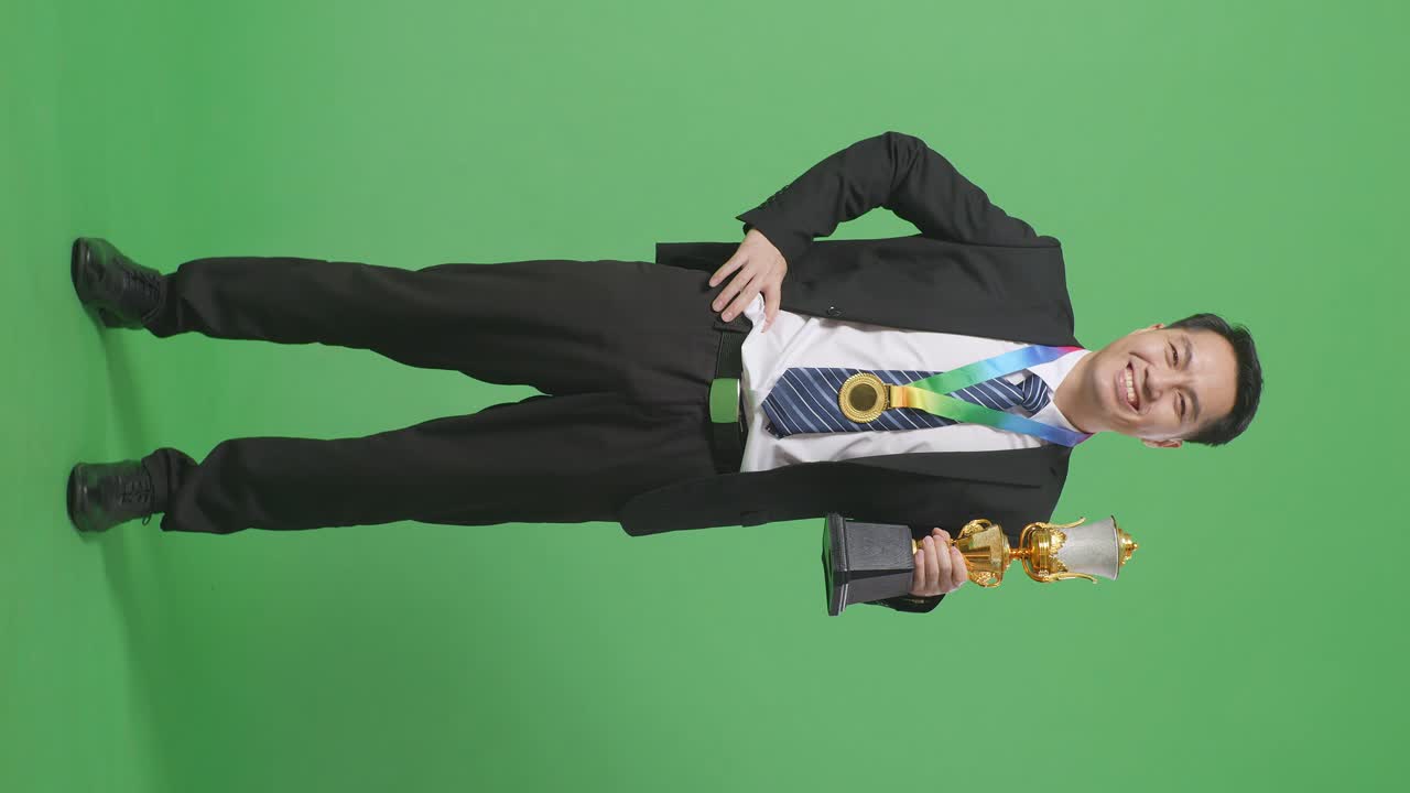 슈트와 타이를 입고 금메달과 트로피를 들고 카메라를 향해 한 팔로 미소 짓는 아시아 사업가 아보가 스튜디오의 녹색 스크린 배경에서 첫 번째 우승자로 포즈를 취하고 있습니다.