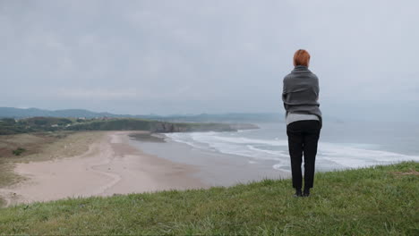 woman standing on a grassy cliff overlooking a serene beach and ocean