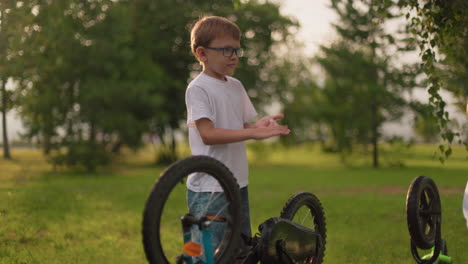 two young siblings are standing next to their upside-down bicycles in a grassy field, the elder brother spins the wheel of his bike while his younger brother watches curiously