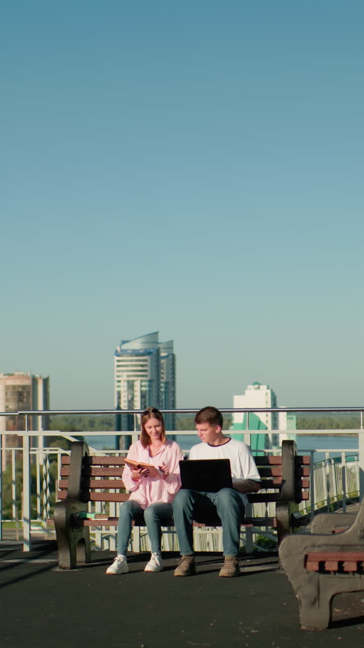 dos estudiantes sentados al aire libre en bancos, un niño escribiendo en una computadora portátil mientras la niña señala algo en su libro, con un telón de fondo urbano de edificios, barandillas y vegetación bajo un cielo azul claro