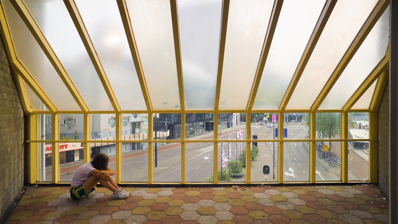 Sad woman sits hugging knees staring out to large windows as town traffic rushes outside, timelapse below frosted glass roof