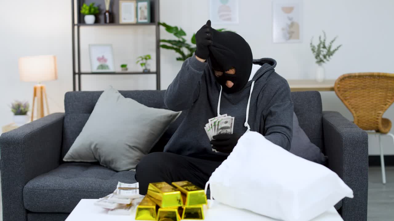 Ein maskierter Mann zählt Geld und Goldbarren auf einem Tisch, bevor er sein Gesicht enthüllt.