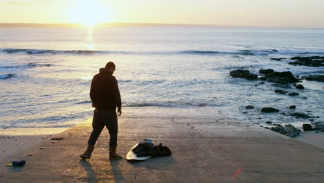 Vista-Trasera-De-Un-Hombre-Adulto-De-Mediana-Edad-Con-Una-Tabla-De-Surf-Parada-En-La-Playa-4k