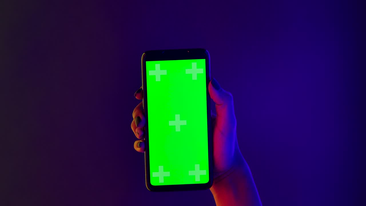 An African American woman holds in her hand a smartphone with a green screen chroma key. Close up of a phone in a vertical position against a background of bright multicolored neon lights. Slow motion