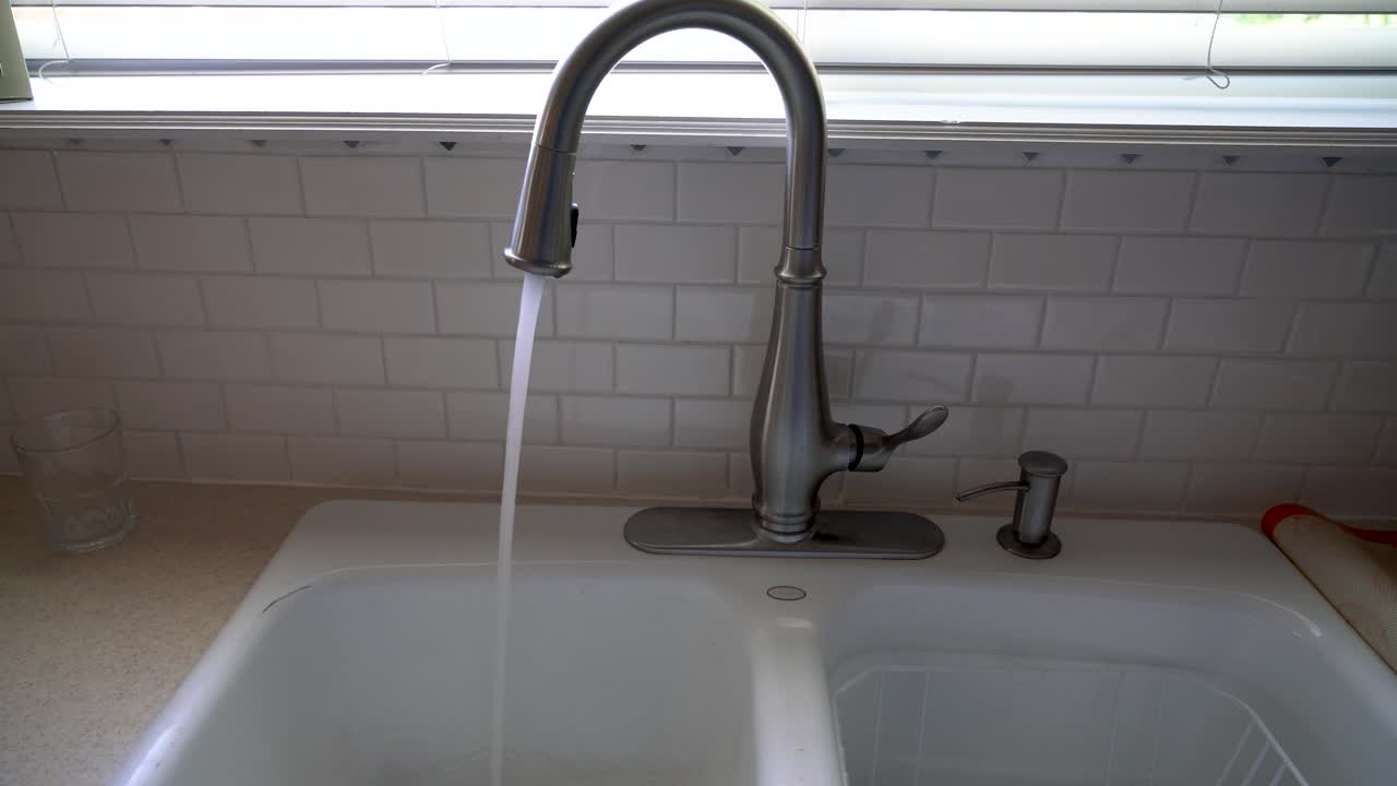 Una toma amplia de agua corriendo de un grifo de acero inoxidable a un fregadero de cocina, frente a un salpicadero de azulejos con formas de diamante azules