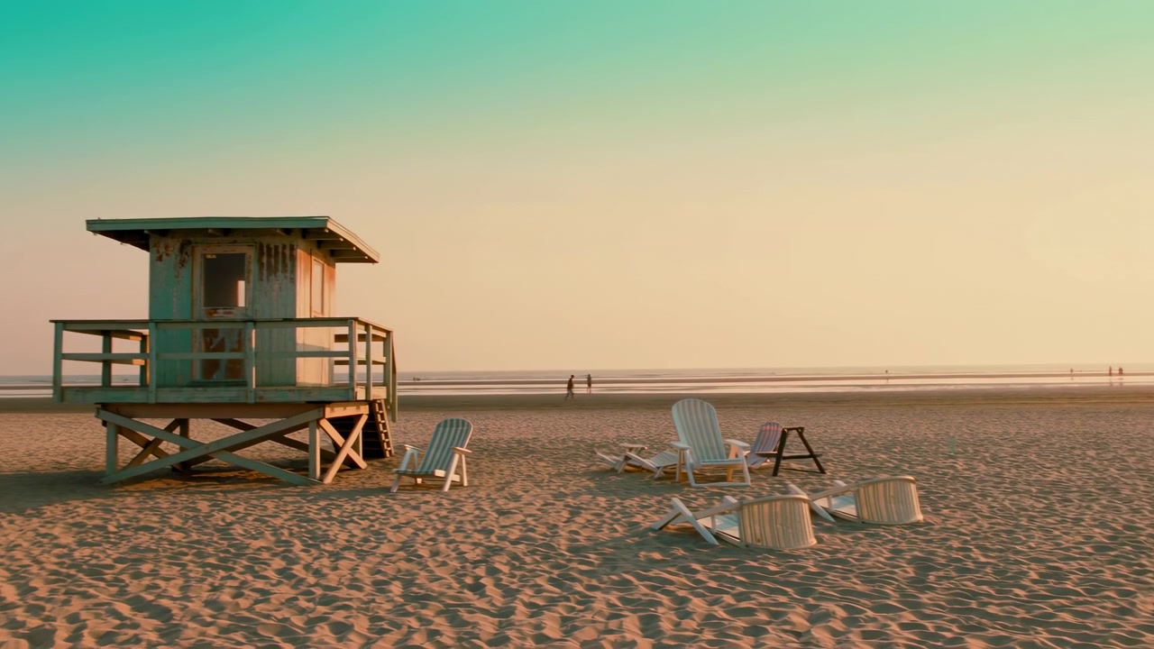 Calm coastal scene showing empty lounge chairs on rippled sand beside small lifeguard hut, with tiny distant people near reflective shoreline under warm golden light and pastel sky