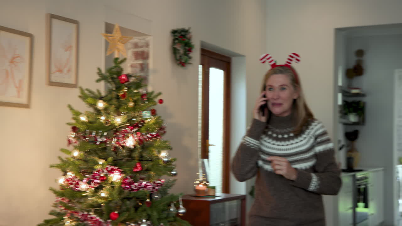Senior woman wearing festive antlers talking on smartphone by Christmas tree