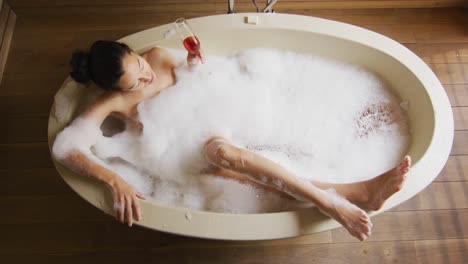 relaxed biracial woman lying in bath with foam and pampering herself with rose petals and wine