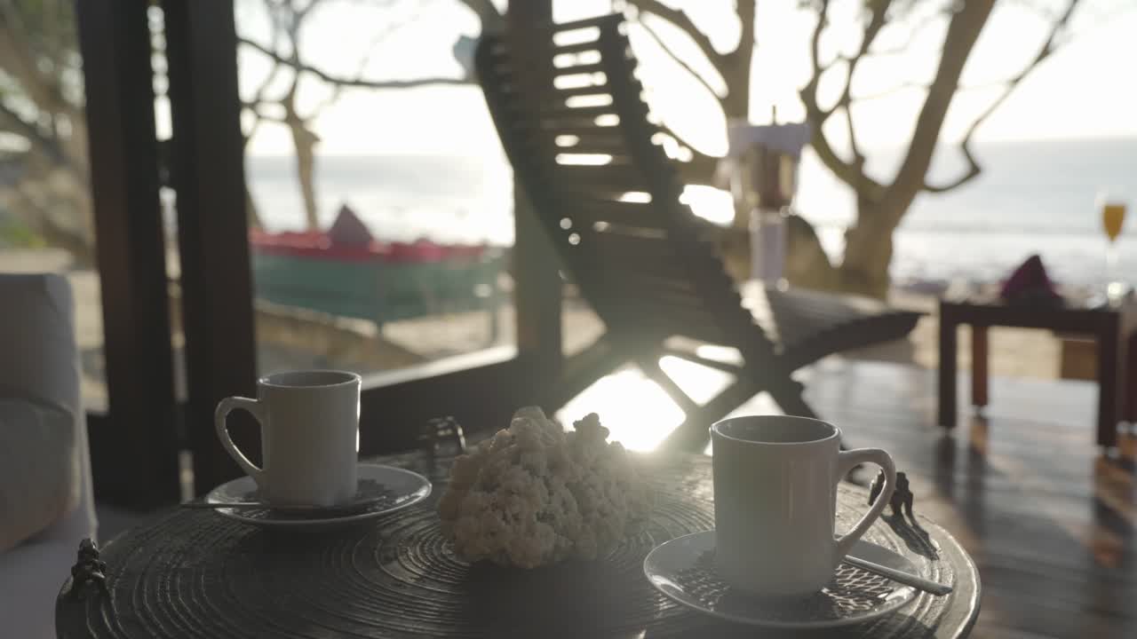 Static shot 2 white steaming mugs on a small table in a tropical luxury villa with the sunny ocean in the distant background