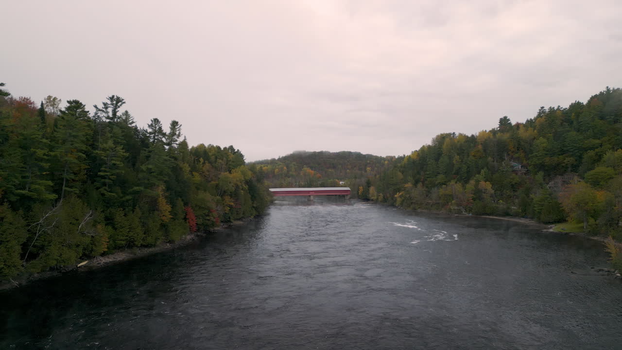 A drone backs away from a red covered bridge to reveal more rolling hill shorelines in the fall season