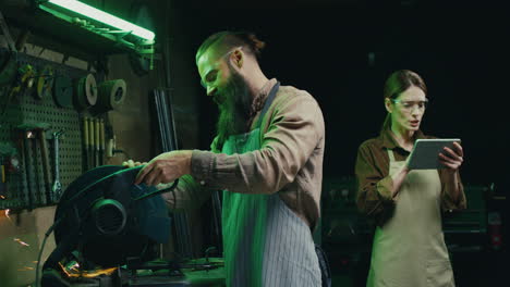 young caucasian female welder in goggles and apron tapping and scrolling on tablet device while watching to her coworker cutting metal on machine in workshop