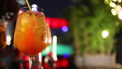 a close-up of a colorful iced cocktail being stirred with a straw at an outdoor night venue in bangkok, with vibrant bokeh lighting
