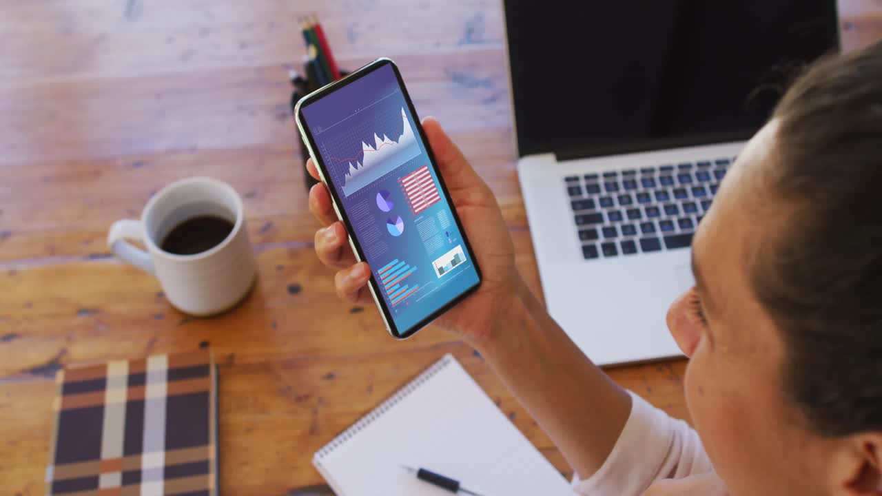 Kvinde af blandet etnicitet sidder ved skrivebord og bruger en smartphone med statistik på skærmen