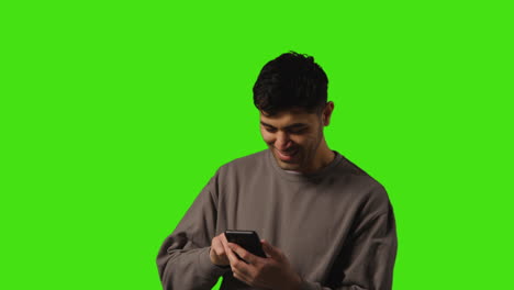 joven mirando un mensaje de texto o navegando en línea en un teléfono móvil de pie contra un fondo de pantalla verde 1