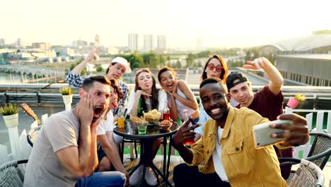 young african american man is holding smartphone and taking selfie with his friends multi-ethnic group holding bottles and glasses enjoying rooftop party.