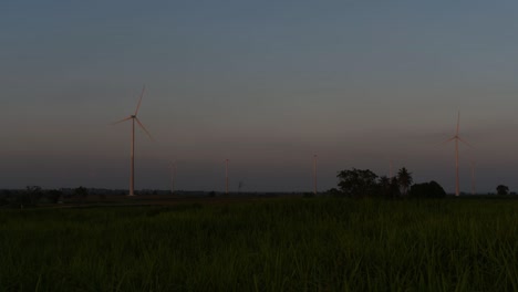 wind turbines shooting out of a farmland while the sun is setting as trucks pass by making clouds of dust, clean alternative energy in thailand and mainland southeast asia