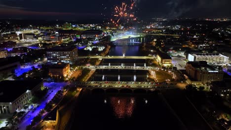 Downtown-Des-Moines,-Iowa-buildings-at-night-with-fireworks-exploding-in-celebration-of-Independence-Day-over-bridges-with-drone-video-tilting-up