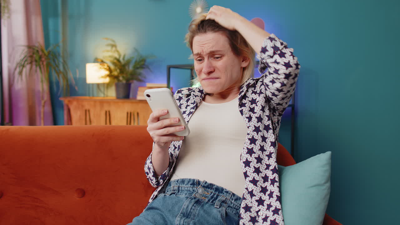 Shocked sad young woman using smartphone reading negative message feels annoyed sitting on sofa