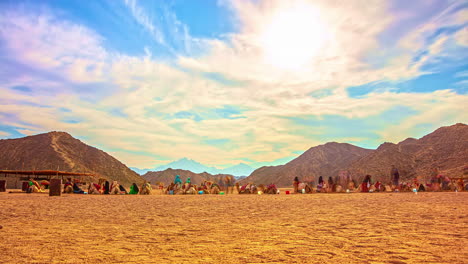 Timelapse-of-people-and-camels-or-dromedaries-resting-in-desert-with-rocky-mountains-in-background-and-clouds-moving-fast-overhead