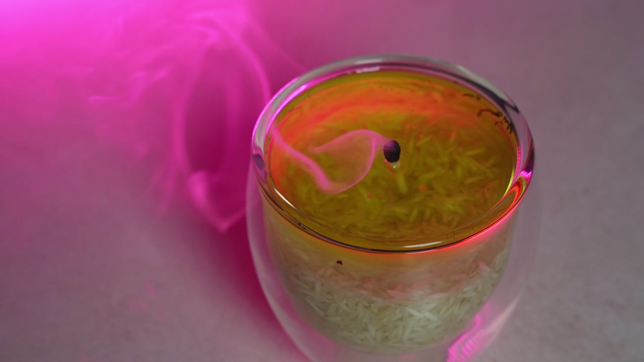 Un primo piano di una candela a olio in vetro fatta a mano che viene spenta su una superficie piana. La fiamma brillante si estingue, lasciando una scia di fumo che sale illuminata da una vivace illuminazione da studio interna rosa