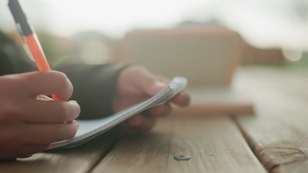 Close up of hand holding pen writing in notebook on wooden table outdoors with blurred open book in background, capturing calm focused study moment in natural light with soft background tones