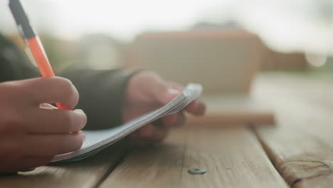 close up of hand holding pen writing in notebook on wooden table outdoors with blurred open book in background, capturing calm focused study moment in natural light with soft background tones