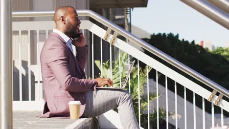 hombre afroamericano en la ciudad sentado en las escaleras con café, usando teléfono inteligente y computadora portátil