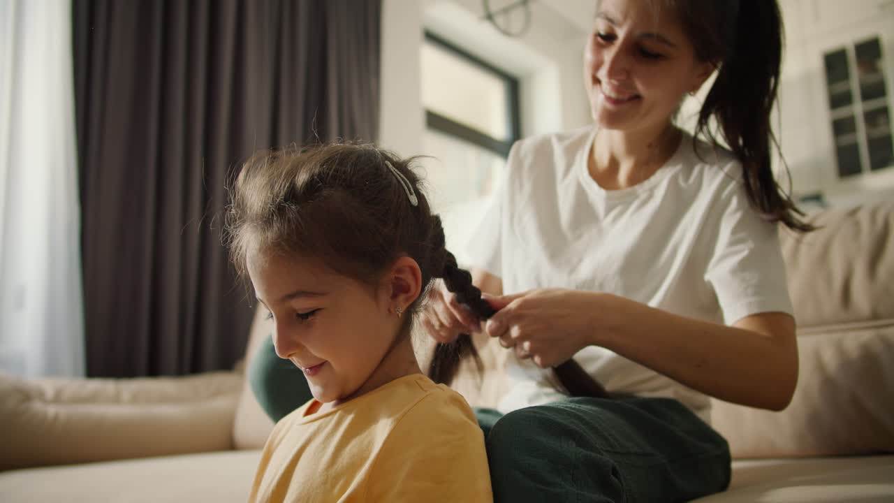 madre morena feliz en una camiseta blanca le da a su alegre hija morena en un vestido amarillo un peinado trenzado mientras está sentada en un marrón claro