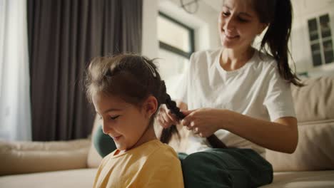 Happy-brunette-mother-in-a-white-T-shirt-gives-her-cheerful-brunette-daughter-in-a-yellow-dress-a-braid-hairstyle-while-sitting-on-a-light-brown