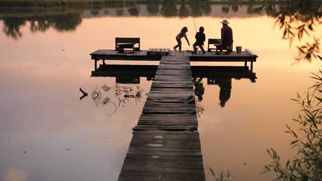 visão traseira distante de um menino adolescente e uma menina sentada com seu avô no cais do lago e pescando juntos