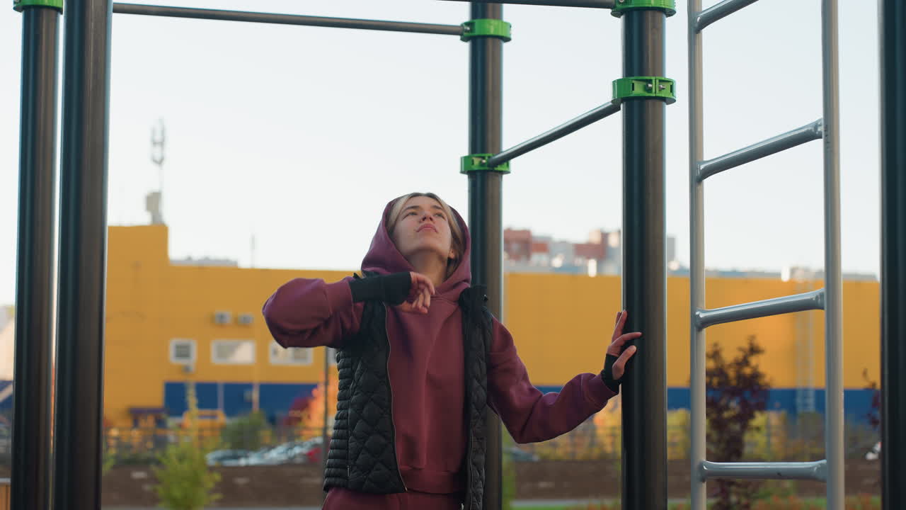 Strength trainer lifting hands to touch horizontal bar while balancing on rubber gym floor wearing burgundy hoodie in outdoor park with bench trees people moving in background before workout