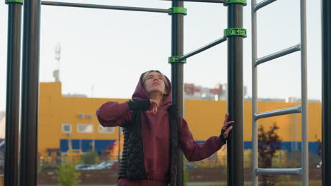 strength trainer lifting hands to touch horizontal bar while balancing on rubber gym floor wearing burgundy hoodie in outdoor park with bench trees people moving in background before workout