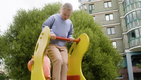 Nahaufnahme-Eines-Kleinen-Mädchens-Mit-Down-Syndrom,-Das-An-Einem-Windigen-Tag-Mit-Anderen-Kindern-Im-Park-Spielt.-Sie-Kommen-Auf-Die-Rutsche