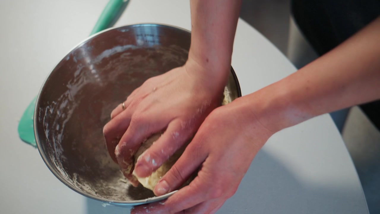 Mãos amassam a massa em uma tigela de metal. Dedos cobertos de farinha. Superfície de mesa branca visível. A cena mostra uma atividade de culinária caseira. Ideal para conteúdo de gastronomia, culinária ou estilo de vida.