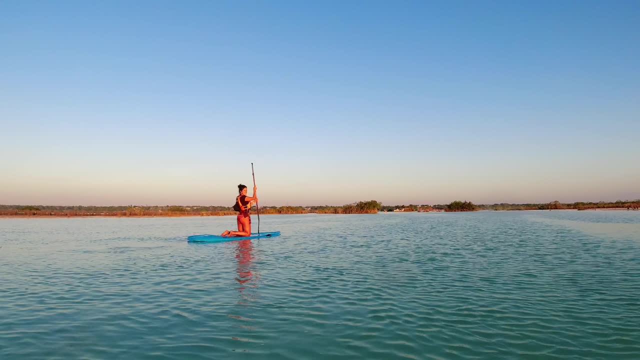 Paddling on a SUP paddle board beautiful young American woman blue lake lagoon still water during sunset
