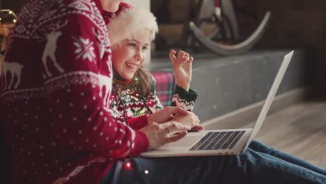 happy little girl in christmas sweater and santa hat sitting on the floor with her mom and looking something on laptop computer 1