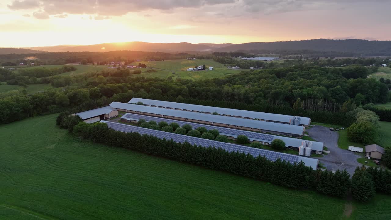 Aerial view of modern farmstead with solar panels on roof of barn building. Golden sunrise behind mountains. Green scenic farm fields and forest trees in suburbia.