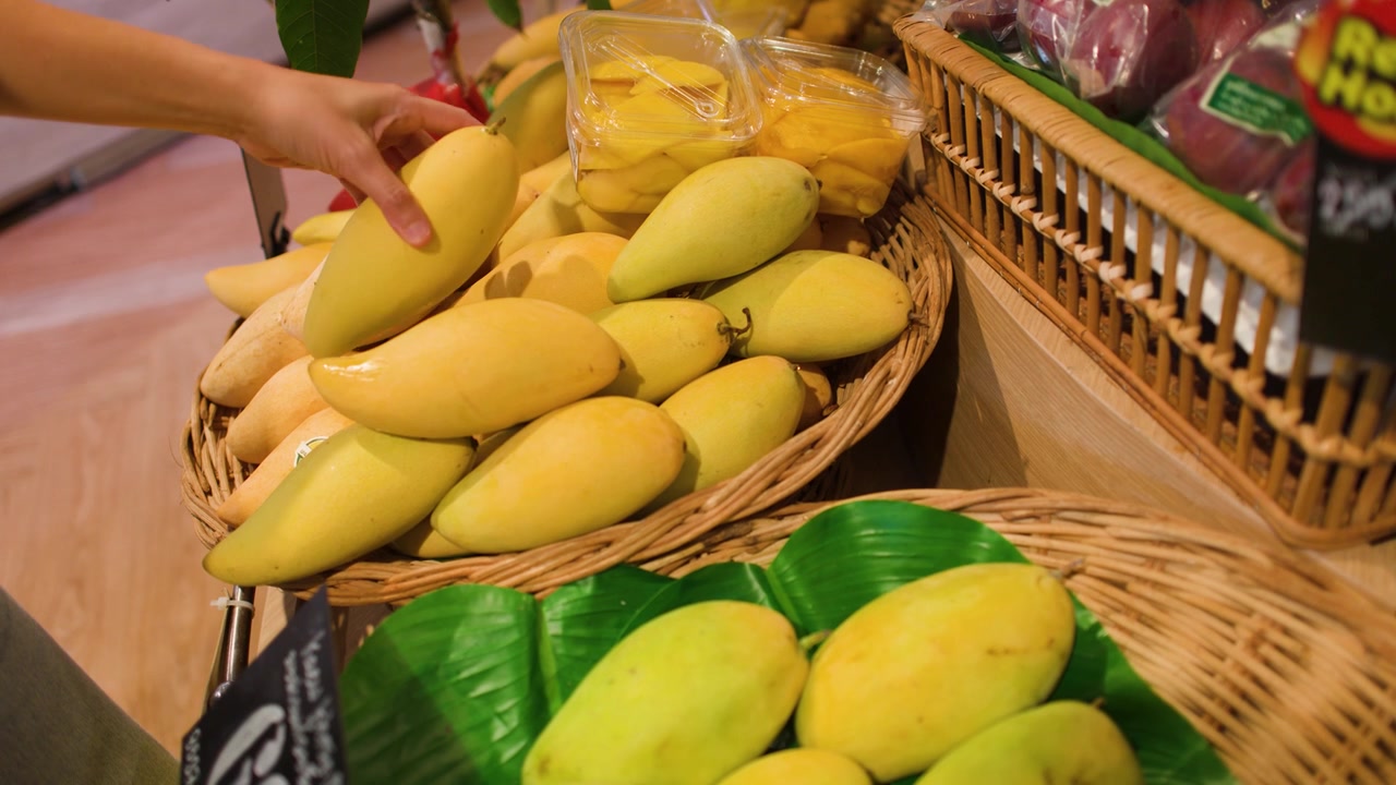 Ręka ostrożnie wybiera i sprawdza dojrzałe żółte mango z ekspozycji w wiklinowym koszu.