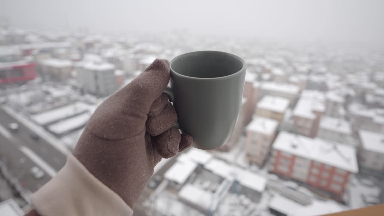 Mano guantata che tiene una tazza su un paesaggio urbano innevato.