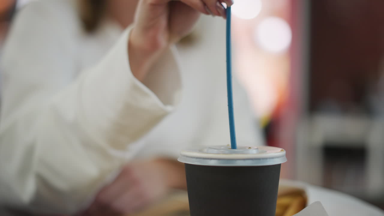 Close-up of hand delicately inserting a blue straw into a takeaway tea cup lid, set against a softly blurred background with vibrant bokeh lights