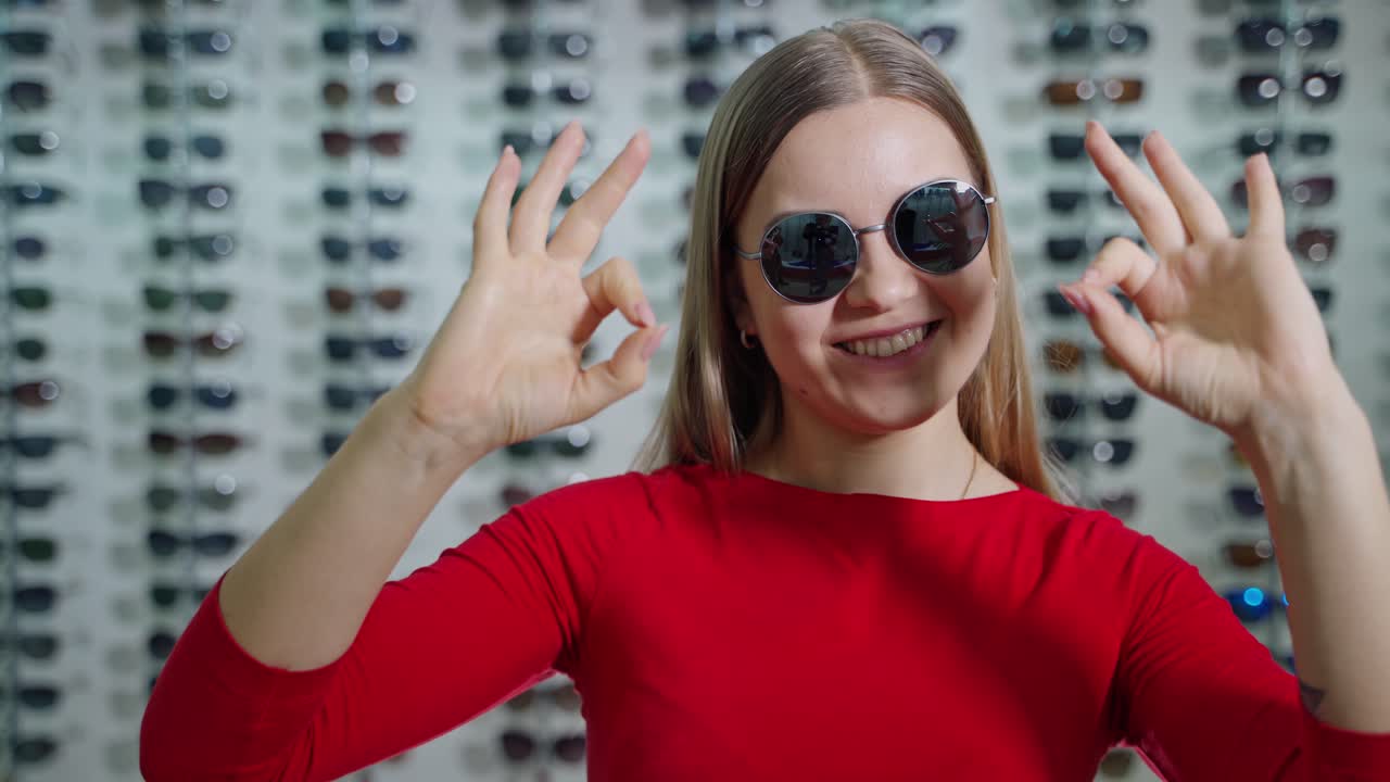 Pretty girl in fashionable sunglasses in the store. Portrait of a lovely girl showing Ok sign by hands on the background of sunglasses at optics.