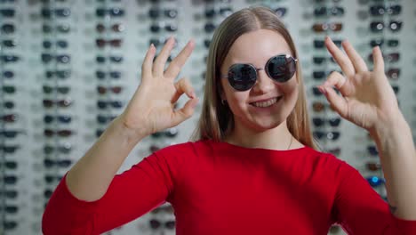 pretty girl in fashionable sunglasses in the store. portrait of a lovely girl showing ok sign by hands on the background of sunglasses at optics.